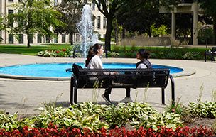 Two students sit on a bench near the Fisher Fountain, with flowers, trees, the fountain, buildings pictured on a sunny, summer day.